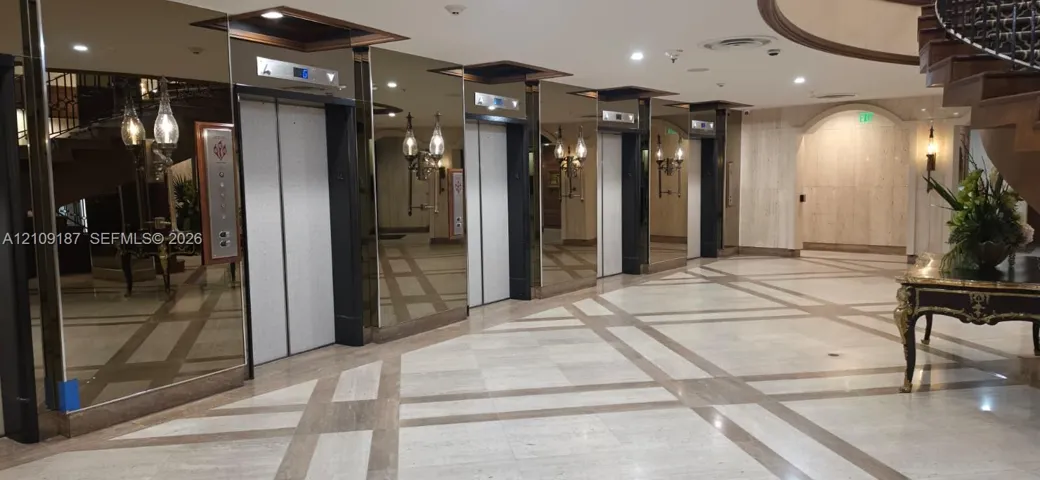 Grand lobby featuring floor-to-ceiling mirrored walls, a grid-patterned stone floor, a sweeping staircase, and multiple elevator banks