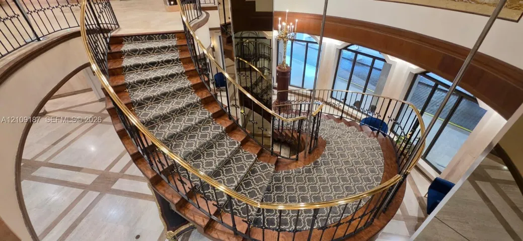 Curved staircase featuring wood-finish treads, custom patterned carpet inlay, wrought iron balusters, and a brass handrail, complemented by stone tile flooring with decorative inlay