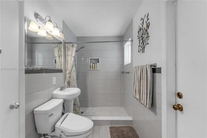 Bathroom featuring full-height gray wall tile, a walk-in shower with a recessed tiled niche, a pedestal sink, and a multi-bulb vanity light fixture