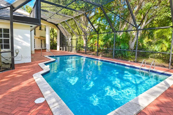 Screened-in pool enclosure featuring a brick paver deck, metal ladder, and perimeter landscaping