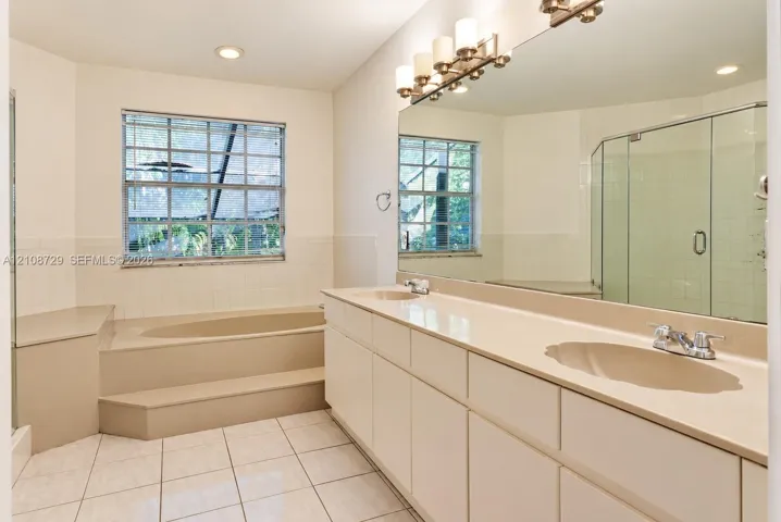 Dual-sink vanity with a solid-surface countertop, soaking tub with a stepped surround, tiled flooring, glass-enclosed shower, and a full-width wall mirror