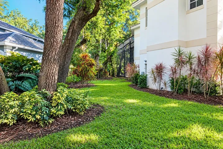 Side yard featuring manicured lawn, established shade trees, mulched garden beds, and screened patio enclosure