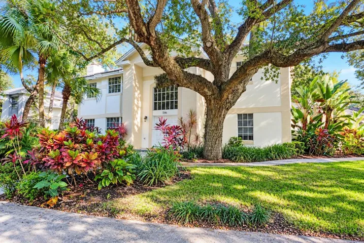 Two-story stucco exterior featuring a grand arched entryway, multi-pane windows, mature oak tree, and landscaped tropical foliage