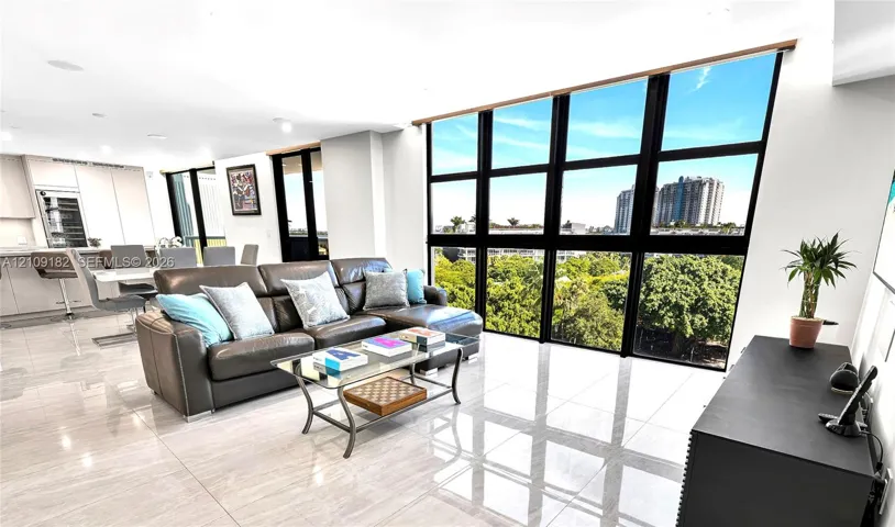 Open-concept living space featuring floor-to-ceiling windows with black mullions, large-format polished tile flooring, recessed lighting, and built-in kitchen cabinetry with integrated stainless steel appliances