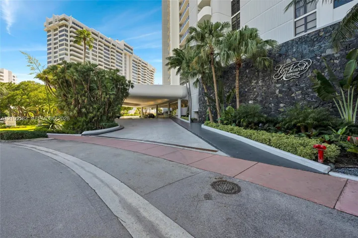 Covered porte-cochère entry with natural stone accent wall, tropical landscaping, and concrete driveway