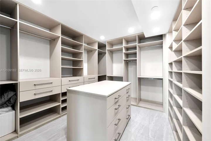 Customized walk-in closet featuring floor-to-ceiling built-in shelving, a central storage island with soft-close drawers, recessed ceiling lighting, and stone-finish flooring