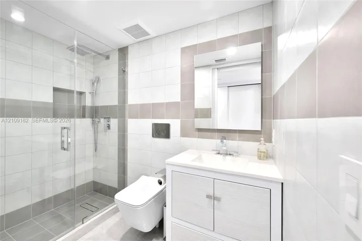 Walk-in glass shower with integrated wall niche and rainfall showerhead, wall-mounted toilet, vanity with light-toned wood-finish cabinetry, and neutral-toned ceramic wall tile