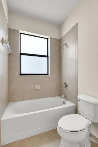 Neutral-toned tile surround, integrated shower-tub combination, rectangular frosted window, and porcelain toilet