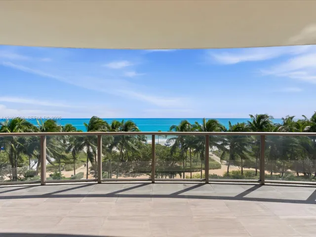 Curved balcony featuring stone tile flooring and a glass panel railing overlooking the oceanfront, sandy beach, and palm trees