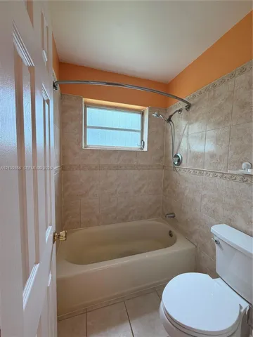 Bathtub with a curved shower rod, wall-mounted hand-held shower fixture, neutral tile surround with decorative inlay, and tile flooring
