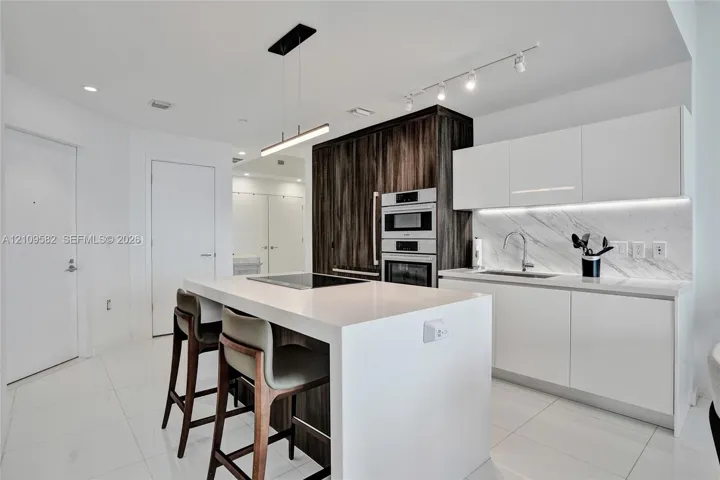 Kitchen featuring a central island with a quartz countertop, flat-panel cabinetry, a built-in wall oven and microwave, stone-finish backsplash, and integrated under-cabinet lighting