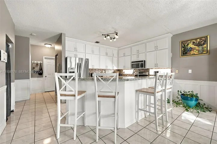 Kitchen featuring white shaker-style cabinetry, a stone countertop, a tile backsplash, and ceramic tile flooring with white wainscoting