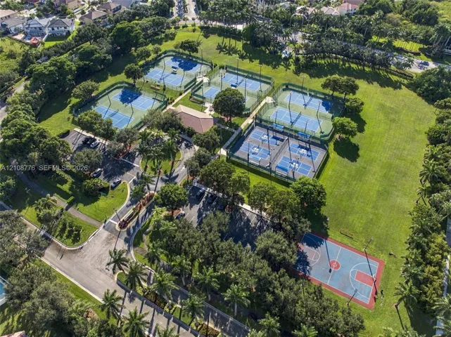 Recreational complex featuring multiple tennis courts, a paved basketball court, asphalt parking areas, and mature trees situated on expansive lawn grounds