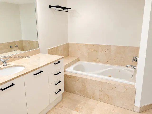 Bathroom featuring a drop-in jetted tub with stone tile surround, stone-topped vanity with undermount sink, white cabinetry with matte black hardware, and a wall-mounted towel rack