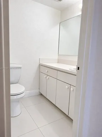 Bathroom featuring white cabinetry, solid-surface countertop with integrated sink, large wall-mounted mirror, white ceramic tile flooring, and standard toilet