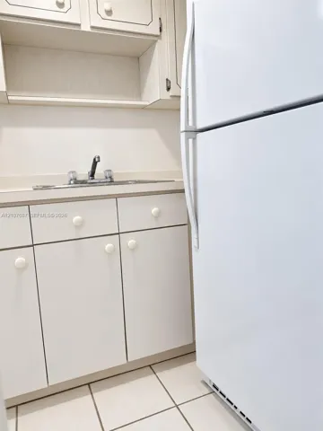 Kitchen area featuring white cabinetry, integrated sink with faucet, tiled flooring, and white refrigerator