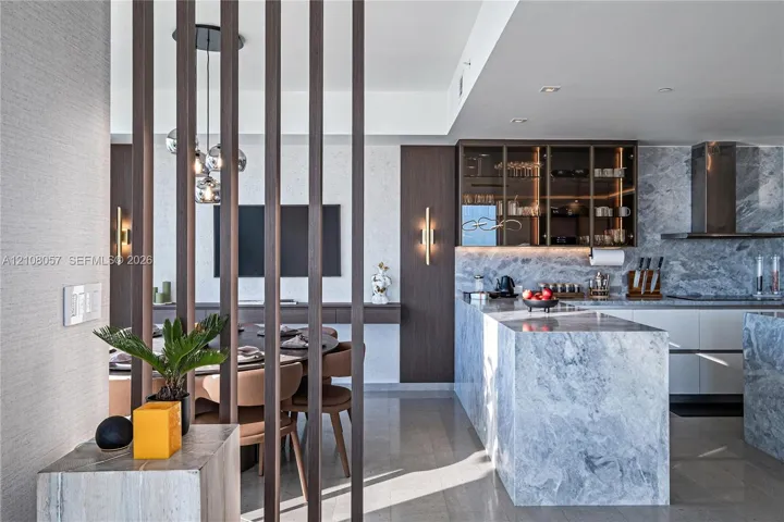 Kitchen featuring stone-clad waterfall island and backsplash, custom cabinetry with glass-front uppers, decorative vertical wood slat room divider, and integrated wall-mounted lighting