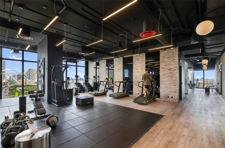 Fitness facility featuring floor-to-ceiling windows, exposed industrial-style black ceilings with integrated lighting, exposed brick accent walls, and dual-tone flooring consisting of square dark tiling and wood-finish planks