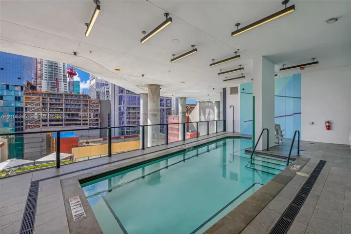 Covered swimming pool featuring linear overhead lighting, structured support columns, and stone-tile decking with integrated drainage