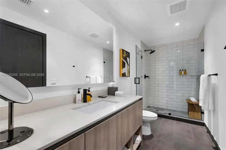 Floating wood-finish vanity with integrated sink and quartz countertop, large backlit mirror, and glass-enclosed shower featuring white subway-style tile and matte black fixtures