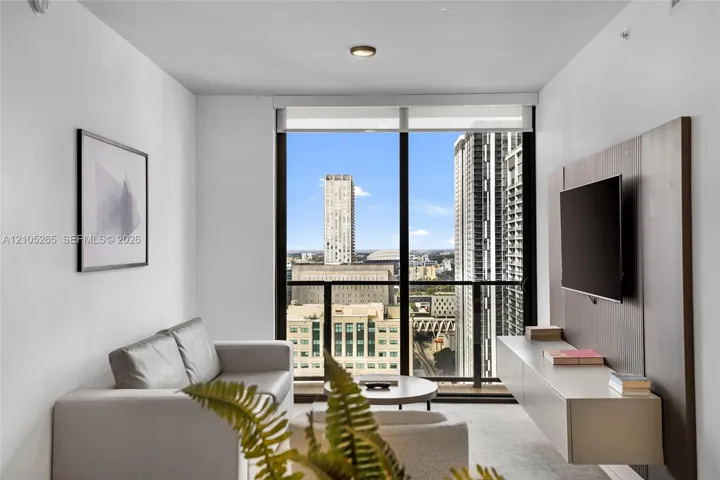 Floor-to-ceiling windows, integrated wall-mounted media console with wood-finish paneling, and private balcony with urban skyline scenery