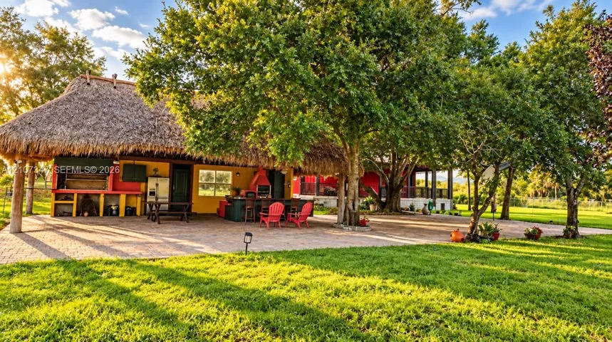 Outdoor living area featuring a thatch-roofed pavilion, stone-paver patio, built-in outdoor kitchen with a grill station, and mature landscaping