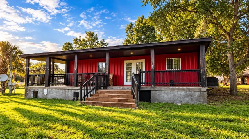 Red metal exterior siding featuring a full-width covered porch, black railing, recessed lighting, and wood-step staircase on a concrete foundation