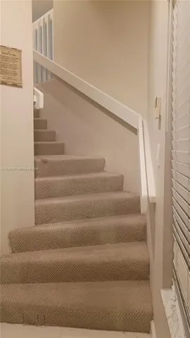 Carpeted staircase featuring a white painted wooden handrail, white vertical balusters, and a window with horizontal blinds