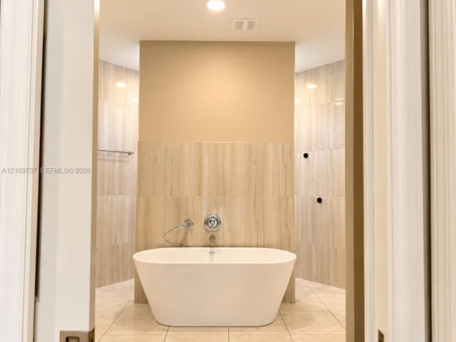 Freestanding white soaking tub with polished chrome wall-mounted faucet and handheld sprayer, set against full-height vertical tile surround and large-format tile flooring