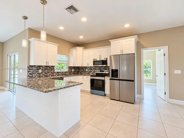 Kitchen featuring white shaker-style cabinetry, stone countertops, stainless steel appliances, a multi-tone tile backsplash, and two glass pendant lights