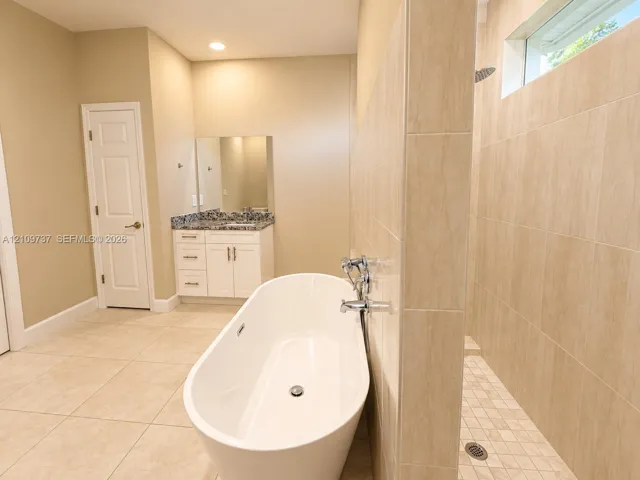 Freestanding soaking tub with floor-mounted faucet, walk-in shower with tiled walls, vanity featuring stone countertop, recessed lighting, and neutral ceramic tile flooring
