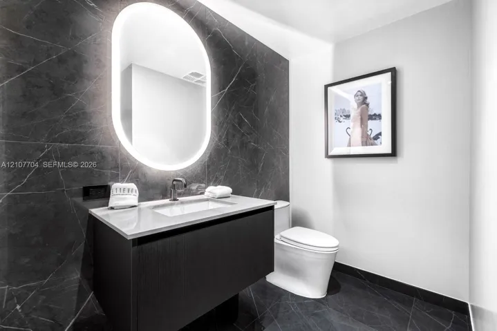 Floating vanity with a light-toned countertop, dark stone-finish wall tile, integrated oval back-lit mirror, and modern wall-mounted faucet