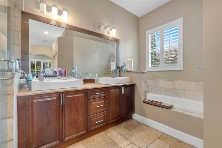 Dual vanity featuring dark wood cabinetry, rectangular vessel sinks, stone-finish countertops, full-width mirror, and two wall-mounted light fixtures, accompanied by a soaking tub with stone tile surround and white louvered shutters