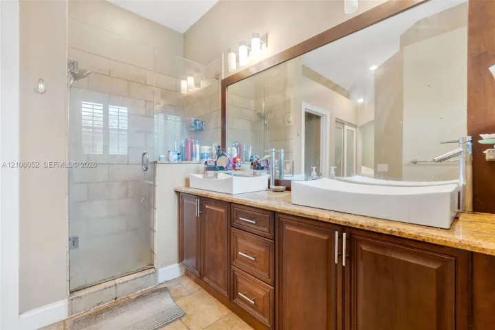 Dual-vanity bathroom featuring a stone-topped wood cabinetry unit, rectangular vessel sinks, polished chrome faucets, a large wall-spanning mirror, and a glass-enclosed shower with tile surround