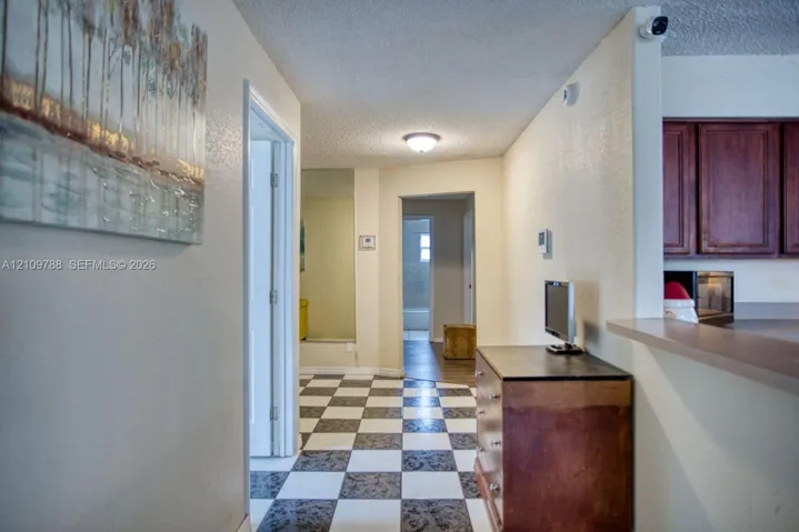 Interior hallway featuring high-contrast checkerboard flooring, a wall-mounted mirrored panel, and a pass-through opening into the kitchen area with dark wood-finish cabinetry