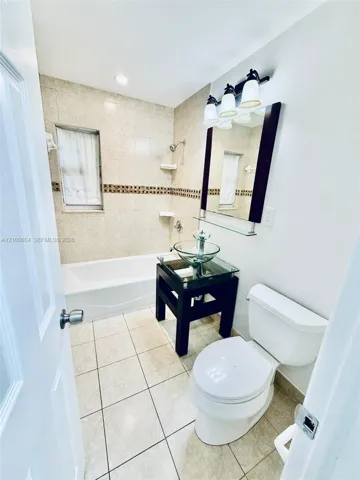 Full bathroom featuring light-toned tile flooring, a bathtub surround with a decorative mosaic tile inlay, a glass vessel sink vanity, and a three-light wall-mounted fixture