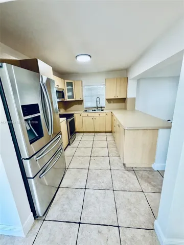 Kitchen featuring light wood cabinetry, stainless steel appliances, neutral solid-surface countertops, dual-basin stainless steel sink, and large-format tile flooring