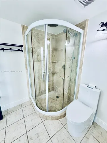 Corner shower enclosure featuring a rounded glass door, integrated stone shelving, and overhead rainfall showerhead, complemented by a dual-flush toilet and large-format tile flooring