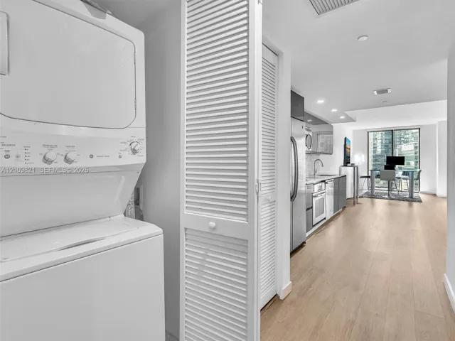 Stacked laundry unit enclosed by white louvered doors, light-toned wood-finish flooring, stainless steel kitchen appliances, and recessed ceiling lighting
