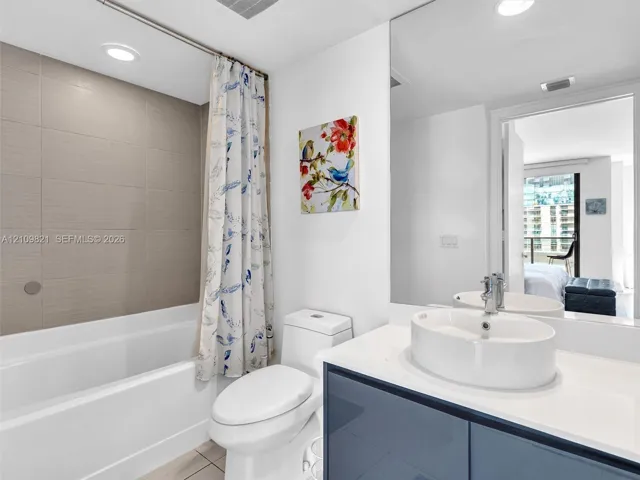 Bathroom featuring a glossy vanity with a solid-surface countertop, an oval vessel sink with a chrome faucet, full-width wall mirror, and a tiled shower surround