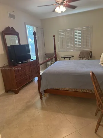 Bedroom featuring large-format ceramic tile flooring, plantation shutters, and a ceiling-mounted light fixture with a fan