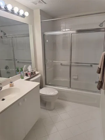 Full-width vanity with an integrated sink, polished chrome light fixture, large wall-mounted mirror, white tile flooring, and a shower-tub combination featuring sliding glass doors