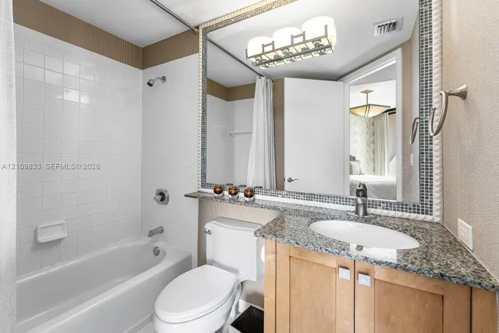 Bathroom featuring a vanity with stone-finish countertop, undermount sink, large mirror with mosaic tile trim, white tile shower surround, and contemporary three-light fixture