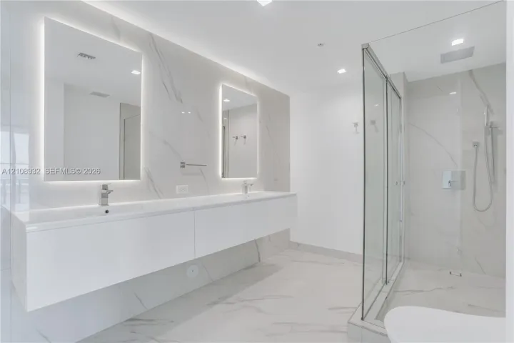 Floating dual vanity with integrated sinks, back-lit mirrors, walk-in glass shower enclosure, and full-height stone-finish wall tiling