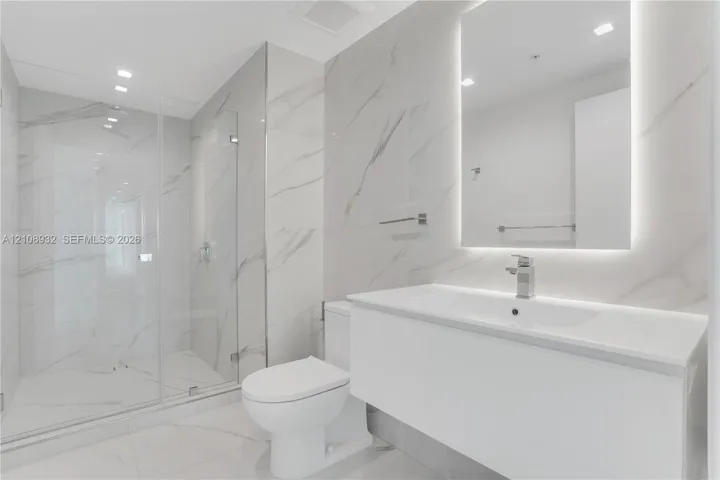 Modern bathroom featuring floor-to-ceiling large-format stone-patterned tile, a glass-enclosed walk-in shower, a white floating vanity with a seamless countertop, and a wall-mounted mirror with integrated backlighting