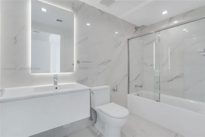 Bathroom featuring large-format stone-patterned wall and floor tile, a modern floating vanity with an integrated sink, a frameless glass tub enclosure, and an LED-backlit mirror