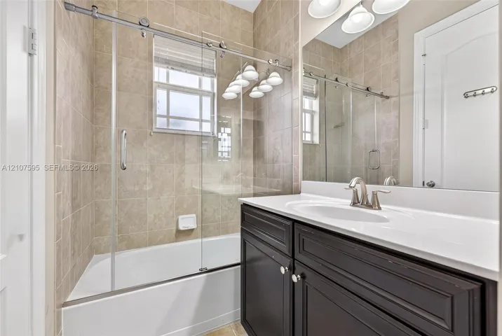 Bathroom featuring a dark-finish vanity with an integrated white countertop and sink, framed glass sliding shower door, and neutral-toned wall tiling
