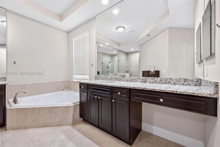 Built-in dark wood vanity featuring a speckled stone countertop, large-scale wall-mounted mirror, corner jetted soaking tub with tile surround, and neutral tile flooring