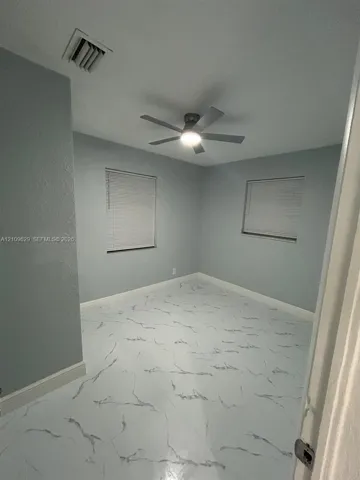 Neutral-toned room featuring large-format tile flooring with marble-patterned veining, dual windows with fitted blinds, and a multi-blade ceiling fan with integrated lighting