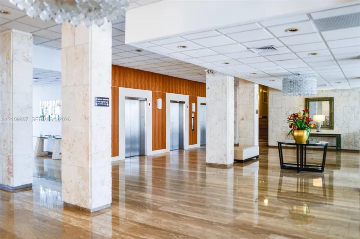 Lobby featuring polished stone flooring, stone-clad structural columns, wood-paneled elevator surround, suspended crystal-style light fixtures, and a recessed drop ceiling